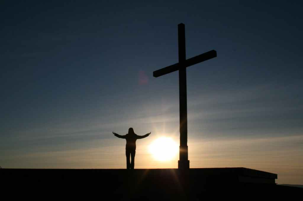 Picture of a cross, a person next to it with arms wide open while the sun is setting. 